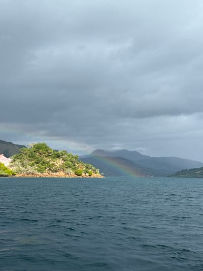 The image shows a view of the sea and land, likely taken near Rinca Village, Indonesia. The sky is overcast with dark grey clouds. A partial rainbow arcs from the sky down to the land, which is covered in green vegetation. The sea is choppy with dark blue water. There are no people visible in the image.
