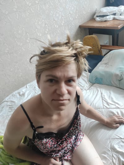 In a dimly lit bedroom in Kostopil, Ukraine, a person with short, spiky blonde hair is lying on a bed covered with a beige duvet. They are wearing a floral print slip dress with black straps. The person is looking directly at the camera with a serious expression. In the background, a bedside table holds a stack of white clothing, and a yellow upholstered chair is visible. The walls are covered with patterned wallpaper. A light green blanket is draped over the edge of the bed on the left side of the frame.