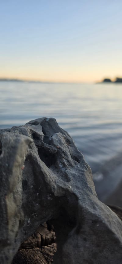 The image depicts a close-up of a weathered, porous rock formation in the foreground. The rock is gray and textured, with several natural openings and irregularities. Behind the rock, a body of water stretches to the horizon, reflecting the soft colors of a twilight sky. The water appears calm with subtle ripples. In the distance, a blurry landmass, possibly a shoreline with faint silhouettes of structures or trees, is visible against the horizon. The sky transitions from a pale blue at the top to a warm, soft yellow-orange glow near the horizon, indicating either sunrise or sunset. There are no people, animals, or any discernible activity in the image. The scene appears to be a natural landscape. The location is stated to be Issyk-Kul, Kyrgyzstan.