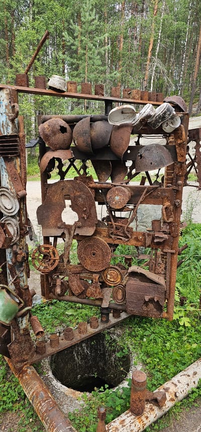This image shows an outdoor scene in Medvezhyegorsk, Russia, featuring an assemblage of rusted metal objects, likely an art installation or monument. The primary subject is a large, rectangular structure constructed from salvaged metal parts, including gears, pipes, bowls, and other industrial fragments, all heavily oxidized and weathered. The structure is set against a backdrop of a forest with predominantly green deciduous and coniferous trees. In the foreground, there is a circular opening or well, surrounded by concrete and encroaching green clover and grass. Additional rusted metal elements, including pipes and cylindrical objects, are present in the foreground, resting on the ground and around the opening. The lighting suggests daylight, with the sun casting some shadows. No people or animals are visible. The overall impression is one of decay and artistic repurposing of discarded materials within a natural, wooded environment.