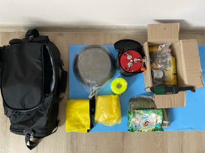 The image displays a flat lay of camping or outdoor gear arranged on a light blue mat. The items are situated on a wooden floor against a white wall. The primary objects include a large black backpack positioned on the left, a covered pot or pan in the center-left, a red portable stove with a black bag, a roll of bright yellow cord, and a box containing various camping supplies such as gas canisters and other equipment. To the right of the pot, there's a green sleeping bag or similar item, and further to the right, a package of baby wipes with a jungle-themed design. A knife with a green handle is also visible, resting on the cardboard box. The scene is an indoor setting, likely a room in a residential building in Bielsko-Biała, Poland, given the context provided. No people are present in the image.