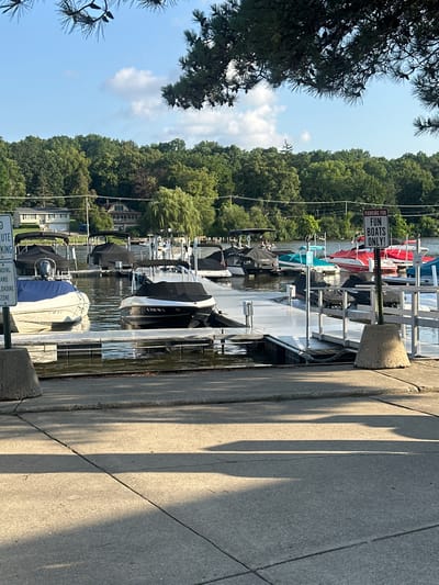 The image captures a scene at a marina on the Fox River in Algonquin, Illinois, United States of America. The foreground shows a concrete walkway with shadows from overhead foliage. Beyond the walkway is a dock with several boats moored. Some boats are covered with tarps, while others are open. The boats vary in size and type, including what appear to be jet skis and smaller motorboats. A sign with the text "PARKING FOR FUN BOATS ONLY!" is visible on a post. In the background, a lush green treeline borders the river, with houses and buildings peeking through the trees. The sky is blue with scattered white clouds. The water of the river is calm, reflecting the surrounding greenery and sky. There are no visible people actively engaged in any activity in the image.