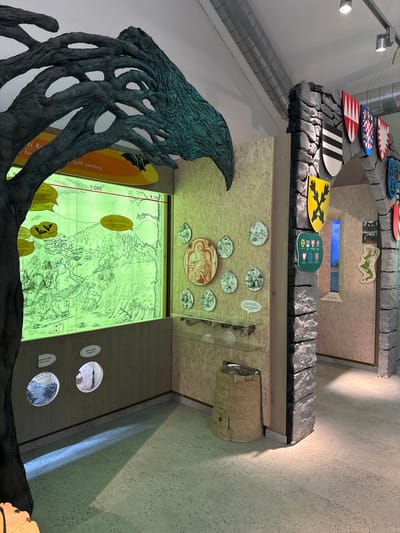 An indoor exhibition space, identified as being in Vavřinec, Czechia, is depicted. The scene features light-colored oriented strand board (OSB) wall panels, a light grey floor, and a white ceiling with exposed silver ventilation ducts and track lighting.
To the left, a large, dark, gnarled sculpture, resembling tree branches and roots with a metallic teal-green sheen on some surfaces, extends upwards and partially across the ceiling, framing a significant portion of the exhibition. Below this sculpture, a large, rectangular, internally illuminated display panel with a green tint is embedded in the OSB wall. This panel exhibits a detailed, historical line-drawing map or landscape featuring figures and structures, overlaid with yellow-orange speech bubbles and bat icons containing text, and a horizontal timeline marked with "10th century" and "1000". Below this main display, two circular cutouts in the wall offer views into internal exhibits, one appearing to contain dark, granular material.
Adjacent to the green display, on the same OSB wall, a cluster of nine circular and oval plaques is arranged. A central oval plaque displays a red-brown illustration of a robed figure, while the surrounding eight smaller circular plaques feature black and white illustrations of various scenes. A small white speech bubble text box is affixed below one of these plaques. Below the plaques, two clear glass shelves are visible; the upper shelf holds several rolled documents or scrolls.
On the floor beneath the glass shelves, a light brown, beehive-shaped cylindrical object with a small, round opening at its base is positioned.
To the right, a faux stone archway, constructed from dark grey, textured material mimicking rough-hewn blocks, serves as an entrance to an adjacent exhibition area. The archway and its immediate surrounds are adorned with multiple heraldic shields of various colors and designs, including black and white stripes, yellow with a black cross, red and white vertical stripes, and white with a red crescent. A green informational sign with smaller shield icons and text is affixed below the yellow shield on the archway's vertical support. Beyond the archway, further OSB walls are visible, along with a vertical light-blue illuminated display and a green map-like cutout on the far wall. No individuals are present in the scene.