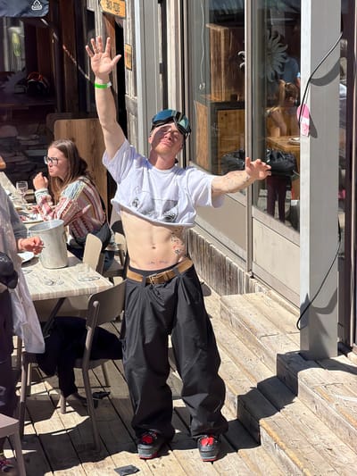 On a sunny day in Saint-Chaffrey, France, an outdoor scene unfolds. A man, shirtless with ski goggles on his head and wearing black ski pants, lies across a wooden deck, posing with outstretched arms. Tattoos are visible on his arms and stomach. A woman with glasses is seated at a table nearby, partially visible and apparently engaged in a conversation. A wine bucket is placed on the table with wine glasses. Reflections can be seen in the glass behind the man, showing a shop window display. The background includes elements of a ski resort environment.