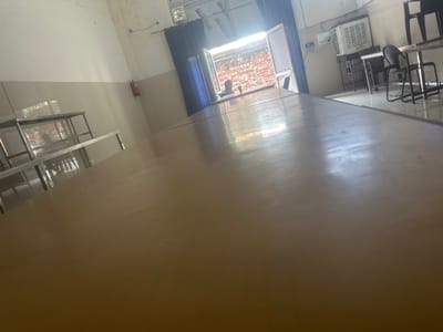 The image depicts an indoor scene, likely a canteen or dining area, in Saunti, India. A long, light-colored table occupies the foreground, its surface reflecting ambient light. In the background, through an open window, a brick wall is visible. A person is seated near the window, facing away from the camera, with a cup on the table in front of them. To the right of the window, a blue curtain hangs. Various furniture, including tables and chairs, are present in the background, along with an air conditioning unit mounted on the wall. A fire extinguisher is mounted on the wall on the left side of the frame. Metal shelving units are also visible. The overall environment appears functional and utilitarian.