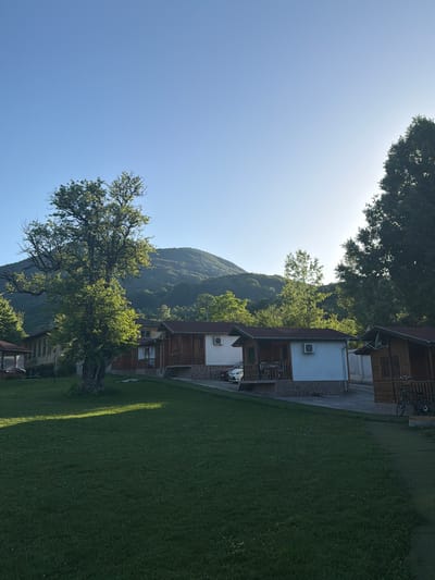 The image depicts a serene setting in Zasele, Bulgaria, featuring a row of small wooden cabins situated in a grassy area. In the foreground, a well-maintained lawn is bathed in sunlight, with a large, mature tree casting a shadow on the grass. Behind the cabins, a forested hill rises, its slopes covered in lush green foliage. The sky is a clear, bright blue, with the sun positioned high and slightly to the right, casting a gentle light over the scene.
The cabins are simple structures, built with wooden walls and dark-colored roofs. Each cabin appears to have a porch or veranda area, and some are equipped with air conditioning units. The cabins are separated by a paved pathway. In front of one of the cabins, a bicycle is parked. A car is also visible parked on the pathway between two of the cabins. The overall atmosphere is peaceful and rural, suggesting a holiday resort or a tranquil residential area.