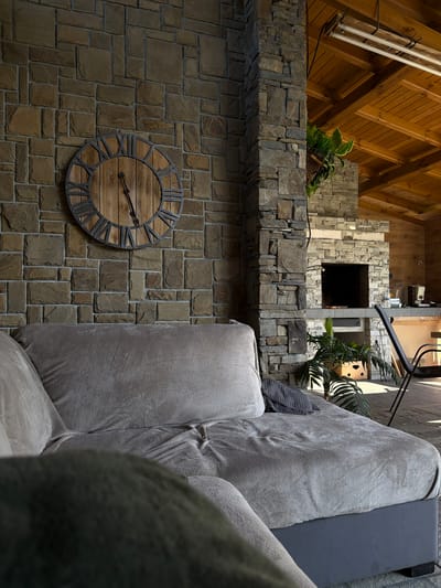 A low-angle indoor shot of a room with a stone wall and wooden ceiling. A large clock with Roman numerals is mounted on the wall to the left. A gray upholstered sofa occupies the right side of the frame, partially obscuring the wall. Above the wall, a stone ledge supports two potted plants. Behind the ledge, a stone structure with a dark opening can be seen, positioned between the ceiling and the wall. The location of this scene is Gorna Rositsa, Bulgaria.