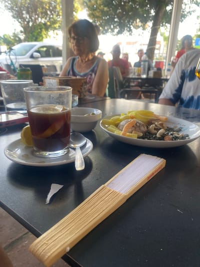 The image captures a close-up view of a table at an outdoor restaurant, likely in San Pedro De Alcántara, Spain, given the presence of a hand-held fan with the inscription "Castellar de la Frontera" which is a town near San Pedro. In the foreground, a wooden-bladed fan lies on a dark, textured surface, possibly a table. To the left, a glass contains a dark beverage with a slice of lemon and ice. It sits on a saucer with a half-eaten lemon wedge and a spoon. Behind the glass, a white bowl holds several green olives. Further back, a plate contains a mix of snacks, including what appear to be dried fruit, nuts, and small cracker-like items, possibly a snack platter or tapa. In the background, blurred figures of people are seated at tables, some appearing to be patrons of the restaurant. The setting is outdoors, with dappled sunlight filtering through trees and overhead awnings, suggesting a warm, daytime environment. A white car is visible in the far left background, partially obscured by foliage. The overall atmosphere is casual and relaxed, indicative of a meal or drinks at a cafe or restaurant.