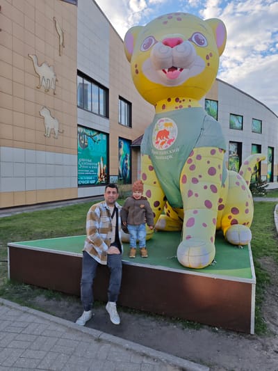 The image shows a man and a young child standing next to a large inflatable leopard at the Yaroslavl Zoo in Russia. The man is sitting on the edge of a raised platform, wearing a plaid shirt, dark pants, and white sneakers. The child, standing next to the inflatable, wears a brown jacket, blue jeans, and a knit hat. The inflatable leopard is yellow with reddish-brown spots and wears a green shirt with the zoo's name and logo written in Russian. In the background, there is a building with animal silhouettes displayed on the facade. A sign with text in Russian is visible near the man. The weather appears to be cloudy or overcast.