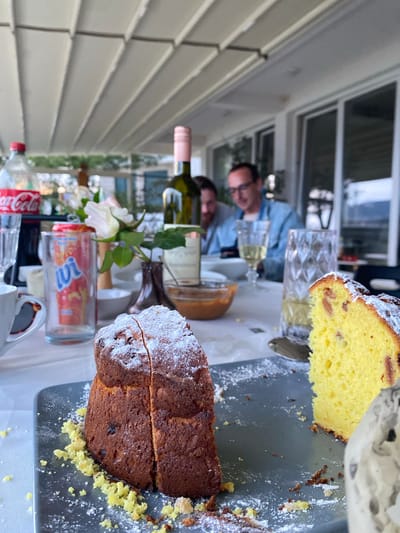 The scene depicts a partially set table with two types of cakes, wine, and soft drinks. One cake is yellow, dusted with sugar, and appears to contain fruit. The other is a darker loaf cake, also dusted with sugar and showing signs of being cut. Beverages include wine in bottles with one having a pink neck seal, and a bottle of Coca-Cola and a bottle of juice. A vase with white flowers is present. In the background, two people are visible seated at the table, one wearing glasses and a blue shirt. The environment suggests an outdoor or semi-outdoor setting, possibly a patio, with a window or opening visible in the background. The city of Trebesin, Montenegro, is not discernible from the given information.