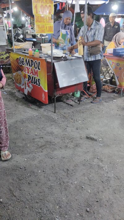 The image shows an outdoor food stall at night in Sambungrejo, Indonesia, likely a market or street vendor area. The stall is decorated with a large banner that reads "SEMPOL AYAM PLUS" and features images of fried chicken and semi-cooked chicken skewers. A yellow sign above the stall displays prices: "1.000" and "SALOTE 1.000", and "TELUR GULUNG 1.000".
A woman wearing a headscarf and an apron is standing behind the counter, appearing to be preparing food. A man next to her, wearing a checkered shirt and a fanny pack, is holding a bottle of yellow liquid, possibly batter or sauce, and seems to be pouring it into a pan. To the right of the man, a person in a black jacket and white head covering is smiling. In the background, more people are visible, and there are other stalls and tents, suggesting a busy commercial area. The ground is covered with gravel and dirt. A motorcycle is parked to the left of the stall. The overall atmosphere suggests a lively night market scene.