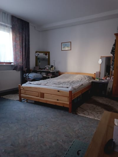 The image depicts a bedroom in what appears to be a domestic setting in Friedberg, Germany. The scene is set during what seems to be late afternoon or early evening, with soft, natural light filtering in from a window on the left, casting long shadows across the room.

The main subject is a wooden bed with a light-colored duvet and a grey blanket neatly spread across it. To the left of the bed, a radiator is visible beneath the window, which is covered by a patterned curtain. Next to the bed, on the right, is a small bedside table with various items on top, including a mirror and what looks like toiletries. Further to the right, a portion of a wooden wardrobe is visible.

In the background, on the wall above the bedside table, a small framed picture hangs. Another, larger mirror with a golden frame is positioned on a dresser or desk unit. A rolled-up blue blanket is placed on the floor near the bedside table, and a patterned rug is visible beneath the bed.

The overall impression is one of a lived-in, somewhat cluttered, but cozy bedroom. There is no discernible activity taking place; the room appears empty and still. No text is visible in the image. The lighting suggests a quiet, possibly restful time of day.