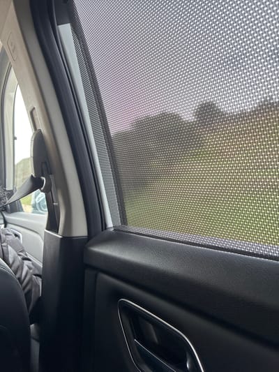 The scene is located inside a vehicle in Krustina, Bulgaria. The primary view is directed toward the passenger-side window, which is covered by a fully extended black mesh sunshade, creating a pixelated effect on the outdoor environment. Through the sunshade, the exterior reveals a green, open field in the midground, with a darker, irregular silhouette, consistent with distant trees or hills, against a light, slightly pink-tinged sky. The vehicle's interior occupies the foreground, featuring a black textured plastic door panel on the right with a partially visible silver-accented door handle. To the left, a light gray B-pillar is visible, equipped with a retracted black seatbelt and buckle. A dark, indeterminate fabric object rests on a seat in the lower-left portion of the frame. No individuals are clearly visible within the image.