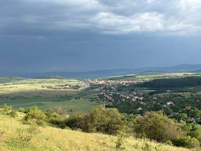 A detailed panoramic landscape photograph captures a sweeping view of a valley containing the town of Dragoman, Bulgaria, observed from a high vantage point. The foreground features a gently sloping terrain covered in dry, golden-green grass and interspersed with small, green deciduous bushes and trees. Descending into the midground, the landscape transitions into broad agricultural fields on the left, displaying varying shades of vibrant green, with patterns of cloud shadow and sunlight illuminating different sections. In the central and right midground, the town of Dragoman is visible, characterized by a dense cluster of residential buildings, many with orange or red-tiled roofs, set amidst an abundance of green trees and vegetation. A prominent multi-lane highway, complete with an elaborate interchange, curves through the valley, passing near the town. In the background, successive layers of green and forested hills recede into the distance, appearing muted under the atmospheric haze. The sky above is largely overcast, dominated by heavy, dark grey clouds, particularly on the left side, indicating possible rainfall. However, brighter, lighter grey clouds and patches of sunlight are visible on the right, casting distinct light and shadow across the landscape. No people, specific actions, or direct interactions are discernible within the frame.