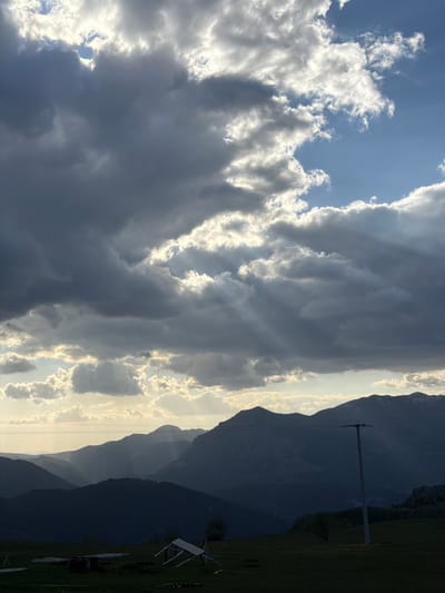 The image shows a landscape of mountains and clouds in Pišče, Montenegro. The left side of the image is dominated by large, heavy clouds, with some patches of blue sky visible. To the right, a silhouette of mountains is seen with sunlight filtering through, indicating the position of the sun. An electrical post is visible on the right side of the mountains. A grassy area is visible at the bottom right, suggesting a field or meadow.