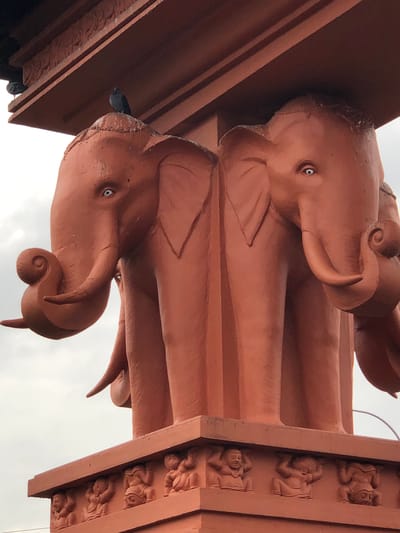 The image shows a close-up view of a red-orange architectural feature, possibly a monument or a building facade, in Sri Jayewardenepura Kotte, Sri Lanka. The central focus is a sculpture depicting what appears to be four elephant heads emerging from a columnar structure. Two elephant heads are prominently visible on the left and right, facing outwards, with their trunks curled. The elephants have bright white eyes with dark pupils. The columnar structure supporting the elephant heads is decorated with relief carvings of small human figures at its base. The sky in the background is overcast. A pigeon is perched on top of one of the elephant heads.