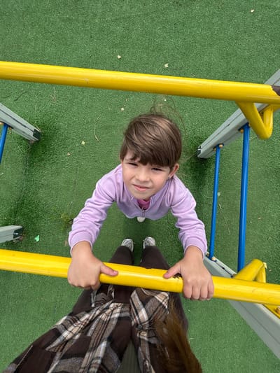 A young girl with brown hair, wearing a light purple hooded jacket and white sneakers, is holding onto a yellow bar on a playground set. She is looking up at the camera with a slight smile. The playground is situated on artificial green turf. A person's legs, wearing brown plaid pants, are visible in the foreground, also holding onto the yellow bar. The perspective is from above, looking down at the girl. The location is identified as Malaya Borshchevka, Russia.