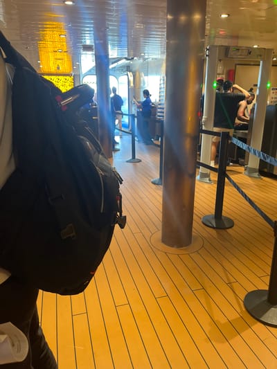 The image shows a line of people waiting to pass through a metal detector at what appears to be a security checkpoint. The floor is made of polished wooden planks with black lines between them. There are metal stanchions with black ropes creating a queue. In the foreground, a person's shoulder and a black backpack are visible. In the background, several people are standing in line, and some are in the process of going through the metal detector. The lighting appears to be artificial, coming from overhead fixtures that reflect off the ceiling. The overall setting suggests an indoor public space, possibly a ferry terminal or an airport. The location is Unknown, United States.