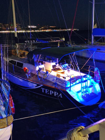 A nighttime view of moored yachts in the city of Krinitsa, Russia. The yachts are illuminated with blue and yellow lights, reflecting on the dark water. The name "TEPPA" is visible on the hull of one of the yachts. In the background, the city skyline is lit with numerous lights, suggesting a lively urban environment. The scene appears to be a serene harbor at night.