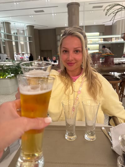 A woman with blonde hair and a light yellow jacket sits at a table indoors, holding a glass of beer up toward the camera. Two empty glasses sit on the table next to her. The background reveals a multi-story interior space, possibly a hotel or resort, with other people seated at tables. The location is likely El Gouna, Egypt, given the user's prompt.