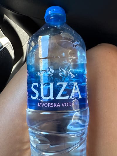 A person is holding a plastic bottle of "SUZA IZVORSKA VODA" (Suza Spring Water). The bottle has a blue cap and a label featuring a mountain landscape and the brand name in large white letters. The person's legs are visible on either side of the bottle, suggesting they are sitting. The background shows a dark car interior. The image appears to have been taken in Bukovac, Montenegro, as indicated by the provided context.