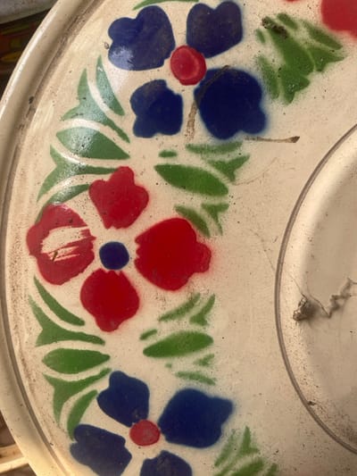 The image displays a close-up, top-down view of a circular, off-white object, consistent with an enamelware plate, tray, or lid. The surface exhibits a painted floral pattern, featuring stenciled four-petaled flowers with alternating color schemes: some are red with a central blue dot, while others are blue with a central red dot. These flowers are interconnected by thin, brown stem-like lines and accompanied by green, elongated leaf shapes. The object's surface is visibly dusty and shows signs of minor wear, including surface abrasions and slight discoloration. A raised, rounded rim is present along the left edge of the object, while a partial concentric circular indentation or raised rim is visible on the right. No individuals or specific actions are depicted. The immediate environment is obscured by the close crop, providing no discernible background details to confirm its precise location within Awgbu, Nigeria.