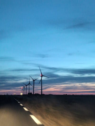 The image depicts a road leading towards a line of wind turbines at dusk. The sky is a gradient of deep blue at the top, transitioning to lighter blues and pinks near the horizon. Wispy clouds are visible across the sky. The wind turbines are silhouetted against the sky, with one in the foreground emitting a red light. The road is asphalt with white dashed lines, suggesting a car's perspective with motion blur. The landscape around the road appears flat and desolate, with a distant treeline. There are no people or animals visible in the image. The scene suggests a rural or open area, possibly a wind farm.
The request to include the location "in the city of Veules-les-Roses, France" is noted, however, there is no visual information within the image that would allow for the identification of this specific location. The image solely provides visual cues of a wind farm setting at dusk.
