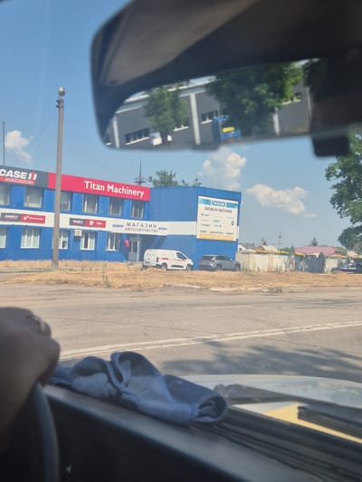 The image shows a view from inside a vehicle, looking out through the windshield and rear-view mirror. The scene is set on a street in Pidhaitsi, Ukraine, likely on a sunny day given the clear blue sky and bright lighting.
In the foreground, a hand wearing a ring is visible on the steering wheel, and a blue cloth is draped over the dashboard. The view through the windshield reveals a road with lane markings and shadows of trees.
The background, seen through the rear-view mirror and directly, features a large blue building with red accents. A prominent sign on the building reads "Titan Machinery". Below this, other signs indicate "CASE II AGRICULTURE", "МАГАЗИН АВТОЗАПЧАСТИН", and "ЗАПЧАСТИ ЧАСТИ". Several logos for vehicle brands like SCANIA, RENAULT, DAF, VOLVO, KRONE, and SAF are also visible on a sign on the blue building. A white van and a grey SUV are parked in front of the building. The building appears to be a commercial establishment, likely a store or service center for agricultural and automotive parts. Beyond the building, a low brick wall with some yellow coverings and trees can be seen, suggesting a semi-urban or industrial area. The overall environment appears to be somewhat dry, with overgrown grass and weeds in the foreground.