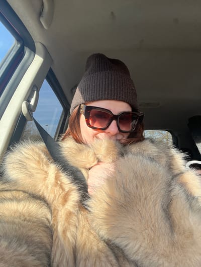 The image displays an individual, appearing to be an adult female, seated within the front passenger compartment of a vehicle during daylight hours. The specific location within Marcq-en-Barœul, France, is not identifiable from the visual content.
The individual wears a dark brown knitted beanie and large, square-framed tortoiseshell-patterned sunglasses, featuring prominent gold-colored "Miu Miu" logos on the temples. Reddish-brown hair is visible beneath the beanie and around the sunglasses. She is dressed in a voluminous, light brown or beige fur coat, possibly fox fur, characterized by varying fur lengths and dark tips on some hairs. A black seatbelt is correctly positioned diagonally across her chest, over the fur coat. Her left hand is partially tucked into the fur, obscuring the lower portion of her face from the mouth down. A subtle upward curve is discernible beneath the sunglasses, consistent with a slight smile.
The vehicle's interior shows a light-colored (off-white or light grey) headliner with visible minor discolorations or marks. A dark red or maroon A-pillar and door frame are visible on the left side, featuring a grey plastic seatbelt guide. Through the car windows, a clear blue sky and bright sunlight are evident. Blurred green foliage and a faint dark vertical pole or structure are visible in the distant background, suggesting an outdoor environment. Strong natural light illuminates the scene, creating distinct highlights and shadows.