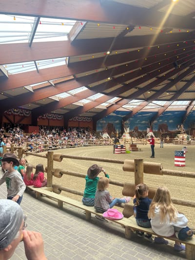 The image depicts an indoor arena with a sandy floor, likely used for equestrian events or shows, located in the city of Collendoorn, Netherlands. A large crowd of spectators, consisting of adults and children, is seated on tiered wooden bleachers on the left side of the frame. The arena is decorated with American-themed elements, including a mural depicting a Western landscape with tepees and cowboys, as well as barrels painted with American flags. In the center of the arena, two performers are visible, one in a red shirt and black pants, and another in a dark shirt and jeans, appearing to be engaging the audience. Several children are seated on a bench in the foreground, facing the arena and watching the show. The arena has a sloped, wooden ceiling with skylights and is adorned with strings of red and yellow lights. The overall atmosphere suggests a lively entertainment event.