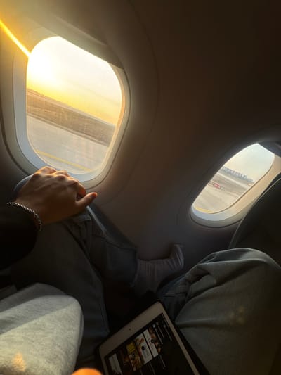 The image depicts the interior of an aircraft cabin, looking outwards through two windows during what appears to be either sunrise or sunset, indicated by the strong golden light. The aircraft is on an airport tarmac or runway, visible outside the windows, with distinct yellow markings on the asphalt. Through the right window, distant airport buildings and structures are discernible, along with a partial marking "08-26" on the ground, suggesting a runway or taxiway designation at Ben Gurion Airport (TLV) in Yehud Monosson, Israel. In the immediate foreground, the left hand of a person is visible, resting on a knee. This hand wears a silver chain bracelet and a dark long-sleeved garment. The person's lower body is clad in light grey sweatpants. To the right, the legs of a second person, wearing light blue jeans, are present. Their right foot, covered in a white sock, is propped up. A white tablet, displaying a media streaming service interface with movie titles like "Kill Bill" and "Forrest Gump," rests on the lap of the person wearing jeans. The scene suggests passengers awaiting departure or after arrival, likely while the aircraft is stationary or taxiing.