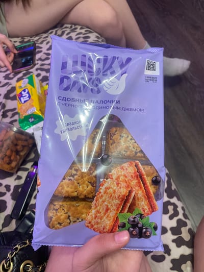 The image shows a close-up view of a purple package of "Lucky Days" cookies with blackcurrant jam. The package is held by a person's hand in the foreground. Inside the transparent window of the package, several cookies are visible, some appearing to be sandwich cookies with jam and a crumble topping, and others showing a more solid layer with jam. There is an illustration of blackcurrant berries and leaves on the lower right side of the package. To the left of the cookie package, a yellow box of "TUC" crackers is partially visible. In the background, out of focus, a person's legs and feet are visible, suggesting they are sitting or lying down. The environment appears to be indoors, possibly a casual setting like a living room or bedroom, with a patterned surface underneath. No specific location in Zmeinyi, Russia, is identifiable from the image.