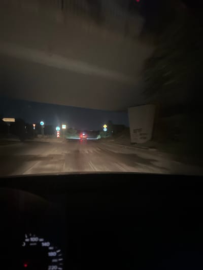 The image depicts a view from inside a vehicle at night, looking out onto a road. The road appears wet, reflecting the streetlights and the headlights of an approaching car in the distance, which has its red taillights illuminated. Several road signs are visible along the sides of the road, indicating directions and regulations. The surrounding environment is dark, with the sky obscured by what appears to be a dark cloud formation or an overhead structure. The dashboard of the vehicle is partially visible in the lower left corner, showing illuminated gauges. The scene is set in Krivina, Bulgaria, suggesting a nighttime drive through the town or its outskirts.
