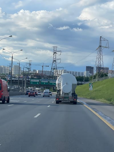 The image shows a multi-lane highway in Novodrozhzhino, Russia. Traffic is present, including a tanker truck. Overhead power lines are visible against a partially cloudy sky. Road signs indicate directions to Moscow-Center, A101 Roslavl, MKAD-West, M4 Kashira, and MKAD-East. A sign also indicates a distance of 400 meters. Buildings, including apartment complexes and construction sites with cranes, are in the background. Streetlights line the side of the highway.