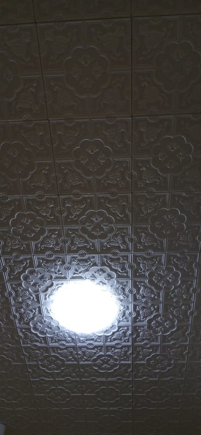The image shows a wall covered in decorative, embossed tiles. The tiles are arranged in a grid pattern and feature an intricate floral motif. A bright, circular light source is prominently featured in the center-right of the image, casting strong highlights and shadows on the textured surface of the tiles. The rest of the wall recedes into shadow, with the embossed patterns still discernible, but less illuminated. The lighting suggests an indoor setting, possibly artificial, given the intense, focused nature of the light. The repetition of the decorative pattern across the multiple tiles creates a sense of depth and texture. There is no visible text in the image, nor any indication of people or specific events. The scene is static and focuses solely on the architectural detail of the tiled wall and the dramatic lighting. The location context of Andijani, Uzbekistan, might suggest a traditional architectural style, though this is not explicitly evident from the image alone.