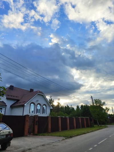 In Цибино, Russia, a white brick house with a brown tiled roof is situated behind a dark brown fence, the fence is composed of corrugated metal panels interspersed with brick pillars. Three arched windows are visible on the front of the house. The house is bordered by a tree line and a street. The sky is filled with dramatic clouds, with patches of sunlight breaking through. A car is parked in the foreground on the left side of the image. Power lines are strung across the sky.