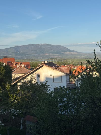 This image captures a serene, sunlit panoramic view of a village or town, identified as Lozno, Bulgaria, set against a dramatic mountainous landscape. The photograph is oriented vertically but is displayed here rotated 90 degrees counter-clockwise.
In the foreground, dense green foliage, including trees and bushes with scattered white blossoms, partially frames the scene. A prominent white house with a reddish-brown tiled roof is visible, featuring a window with a closed roller shutter and another smaller, square window. The warm sunlight creates distinct shadows on the house, suggesting it is either morning or late afternoon. Another building with a similar tiled roof is partially seen just below this house.
The midground expands into a wide valley that appears to be cultivated, possibly with agricultural fields or vineyards, and contains scattered buildings with tiled roofs. Hints of distant infrastructure, such as power lines or roads, can be faintly discerned running through the valley.
The background is dominated by an extensive mountain range, appearing somewhat hazy due to distance. Above the mountains, the sky is a clear, bright blue with a few soft, wispy white clouds.
No people or specific activities are visible, presenting a tranquil and scenic rural landscape. There is no discernible text within the image.
