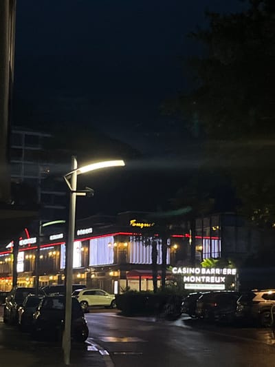 The image presents a nighttime scene in Montreux, Switzerland, specifically depicting the Casino Barrière Montreux complex. The foreground features a wet asphalt street, indicating recent precipitation, with a modern, curved streetlamp prominently positioned in the left-center. A line of dark-colored vehicles is parked along the left curb. In the midground, the multi-story Casino Barrière building is illuminated, showcasing a facade with vertical white light panels and accented by red neon lighting along its upper edges. Visible signage includes "Fouquet's" and a large "CASINO BARRIERE MONTREUX" sign. Palm trees are situated in front of the casino building. On the right side of the street, additional dark-colored vehicles are parked. The sky is dark, consistent with nocturnal conditions. No individuals are visible.