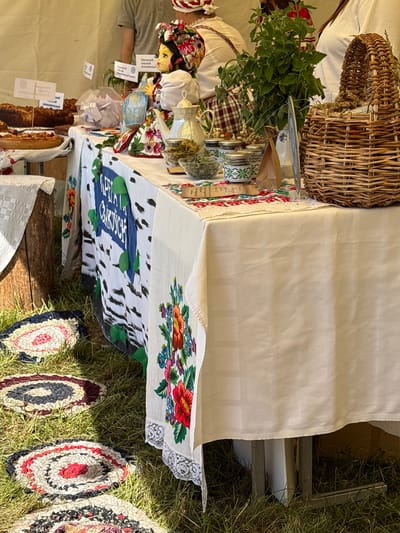 A close-up, eye-level shot captures a market stall with various handmade items and baked goods, possibly at an outdoor festival in Вяйнянскі сельскі Савет, Belarus. In the foreground, several circular, braided rugs made of fabric scraps are laid out on the grass. A white tablecloth adorned with intricate floral embroidery covers the stall's table.
On the table, a traditional doll wearing a colorful headdress and dress sits prominently, holding a blue and green object. Nearby, a silver teapot and several glass jars filled with dried herbs are displayed. Behind these, a woven basket overflows with dried greenery. To the left, portions of baked goods, possibly bread and pies, are visible. The stall is set up outdoors, with a tent-like structure providing shade in the background. The overall atmosphere suggests a local craft fair or market.