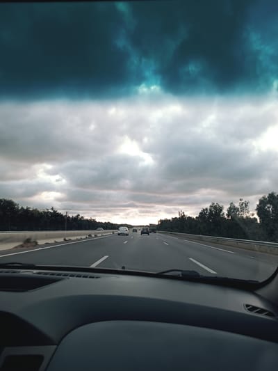 The image shows a view from inside a car driving on a highway. The dashboard is visible in the foreground. The highway has multiple lanes, with other cars visible ahead. The sky is overcast with dark clouds. Trees line the sides of the highway. The location may be on a highway near Ameur, Morocco based on the user's information.