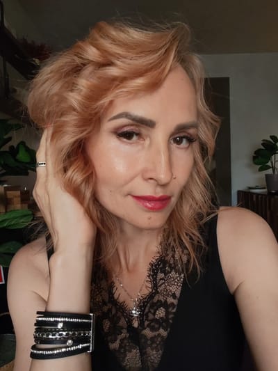 A close-up, head-and-shoulders, outdoor photograph shows a woman with auburn, wavy hair. She has fair skin with a light dusting of freckles and is wearing makeup, including eyeshadow, eyeliner, mascara, and red lipstick. Her left hand is raised to her hair, and she is wearing a ring with three stones on her ring finger. She is also wearing a black top with lace detailing and a silver necklace with a small crown pendant. A black leather bracelet with silver accents and rhinestones is on her left wrist. The background is blurred, but suggests an indoor setting with some greenery and wooden furniture. The image is a selfie, as indicated by the angle and framing. There is no indication of the specific location in Faliukovo, Russia.