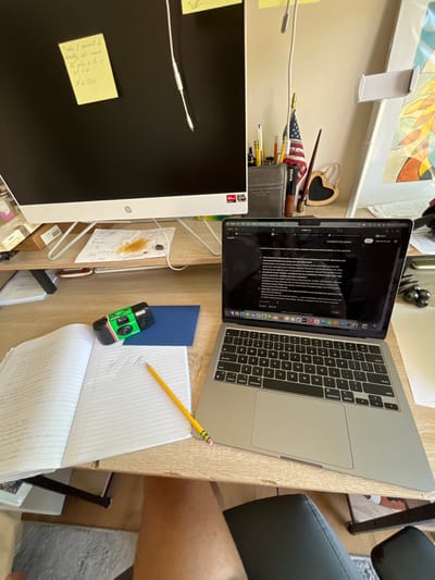 This image depicts an indoor desk workspace, presumably located within a residence in Rancho Palos Verdes, United States.
The primary subject is a light-colored wooden desk with a raised shelf. On the shelf, a large, white HP computer monitor stands prominently, displaying a black screen. A yellow sticky note is affixed to the monitor's upper left, bearing handwritten text. A white audio cable hangs centrally from the top of the monitor. To the right of the monitor, a dark, cylindrical container holds multiple pens and pencils, along with a small American flag and a brown-handled brush or pen. A heart-shaped wooden ornament hangs nearby. Directly beneath the monitor on the shelf, a piece of white paper is stained with a large, irregular brown liquid mark, suggestive of a spilled beverage. A small, dark, smooth object, possibly a stone or bead, rests on this paper.
On the main desk surface, to the right, a silver Apple MacBook laptop is open and powered on, displaying a webpage with dark mode enabled, containing multiple lines of text, seemingly a question-and-answer interface or document. To the left of the laptop, an open, spiral-bound notebook with ruled lines is visible. The left page contains densely written text, while the right page shows a few larger, handwritten words, appearing to be "Hi All." A yellow wooden pencil with a pink eraser is laid diagonally across the open notebook. To the right of the notebook and behind the pencil, a green disposable camera with a white and red striped label is positioned. A dark blue booklet or folder lies partially beneath the camera.
The lower right quadrant of the image shows the upper thigh and knee of a person's right leg, indicating someone is seated at the desk. A black office chair armrest is partially visible directly below the desk. A light grey rug or carpet is partially visible on the floor beneath the desk. The lighting suggests natural light from an unseen source, illuminating the workspace.
