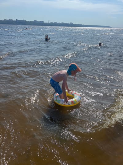 In a body of water, a young boy wearing an orange and blue patterned sun hat and blue and white striped swim trunks stands in shallow water, holding onto a colorful inflatable ring with fruit patterns. He is facing away from the camera, looking towards the water. The water is choppy with sunlight reflecting off the surface. In the background, a distant city skyline with buildings is visible across the water under a clear blue sky. Several other people are in the water, one on a small boat in the middle distance, and others swimming or partially submerged further away. The scene appears to be a sunny day at a waterfront location, potentially a beach or riverbank.