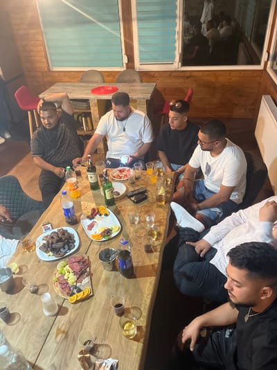 The image depicts an indoor social gathering of approximately seven to eight adult males around a large, light-colored wooden table, in what appears to be a private residence or establishment in Bersin, Bulgaria. The room features wooden paneling on the walls and a visible window with light blue horizontal blinds; this window reflects an interior scene showing additional individuals.
Several men are seated around the table. From left to right:
- One man in a dark patterned t-shirt is seated with his hand behind his head.
- Next to him, a man in a white t-shirt with a dark pendant necklace is looking down at a mobile phone.
- Further right, a man in a dark t-shirt with sunglasses positioned on his head is seated.
- Adjacent to him, a man wearing a white t-shirt, glasses, and denim shorts with rips is seated with his legs crossed, displaying white sneakers.
- At the bottom right, another man in a black t-shirt with a silver chain is visible, looking downwards.
- Partially visible on the left are a man in a white t-shirt and the arm of another individual.
The table is laden with food and beverages. Visible items include:
- Two bottles of Jameson Irish Whiskey.
- Bottles of orange soda, clear water, and a dark carbonated drink.
- Numerous glasses containing amber-colored liquid, some with ice and lemon slices.
- Multiple empty or partially used plastic cups.
- A metal ice bucket.
- A white platter holding dark, chocolate-colored pastries or cookies.
- Another white platter with an arrangement of sliced fruit (strawberries, blueberries, oranges, lemons) and cubes of cheese, along with what appears to be a creamy dip or soft cheese.
- A small wooden serving board presenting cured meats (salami, ham), cheese slices, and grapes.
- A small white bowl containing a red salad, possibly tomatoes and onions.
- Additional items on the table include a cigarette pack, an ashtray, and mobile phones.
In the background, a black electric kettle is visible. A red chair is present on the left side, and two upholstered chairs are visible behind the main table in the upper background, where a small red plate rests on another table. The overall environment suggests a casual and relaxed social event.