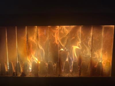 This image depicts a close-up, frontal view of a contained fire burning behind a transparent barrier, observed in Bogdanci, North Macedonia. The primary subject is a dynamic combustion event, characterized by orange and yellow flames, with hints of blue at the base, visible through a pane of glass. This glass barrier appears segmented into multiple vertical panels, exhibiting a light film of residue, possibly soot or dust, which is more concentrated on the lower portions. Below the main flame activity, approximately a dozen dark, conical or ridged structures, consistent with a grate or firebox lining, are visible, also partially obscured by ash or combustion byproducts. The area above and immediately below the visible firebox elements is dark, suggesting an enclosed interior environment with the fire as the dominant light source. No individuals or external objects are visible in the scene.
