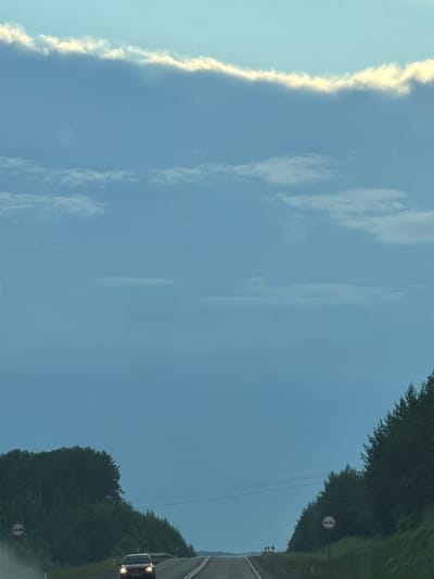 The image captures a road in the city of Karagay, Russia, under a vast, pale blue sky. A thin, luminous band of clouds stretches horizontally across the upper portion of the frame, catching the light. Below this, the sky is a uniform blue, with faint, wispy cloud formations scattered at various altitudes.
The road, paved and appearing to be a two-lane highway, curves gently into the distance. On either side of the road, dense green trees form a natural canopy, creating a sense of enclosure. The trees on the left are closer to the road and form a thick forest. On the right, the trees are also dense, with their branches reaching towards the road.
A red car is visible on the road, moving towards the viewer and emitting light from its headlights. Further down the road, in the distance, a group of small figures, presumably people, can be seen. There are two traffic signs visible: one on the left side of the road, indicating no overtaking, and another on the right side with the same prohibitory symbol. The overall scene suggests a journey through a natural, somewhat remote landscape during daylight hours, possibly late afternoon or early evening given the soft lighting.
