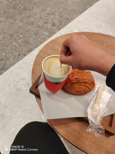 A person's hand stirs a latte in a white paper cup with a red sleeve, alongside a pastry and a wrapped wooden utensil. The coffee and pastry sit on a piece of white paper on a round wooden table. The background shows a gray, speckled surface. The person is wearing black clothing. "HONOR 70 54MP Matrix Camera" appears in the bottom left corner of the frame.