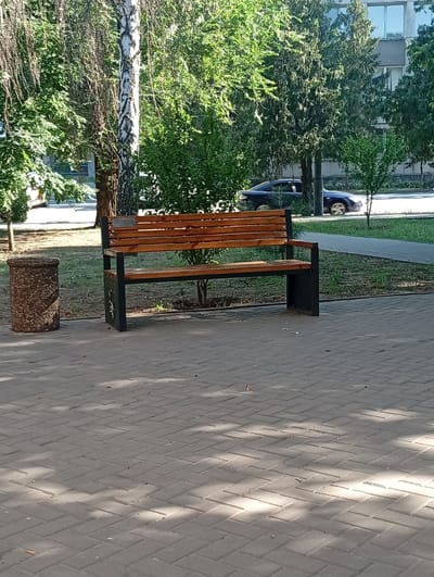 The image depicts a public area in the city of Pokrov, Ukraine, featuring a paved walkway made of interlocking grey bricks. To the left, a cylindrical, rough-textured waste bin stands near a birch tree with distinctive black and white bark. In the center of the frame is a wooden park bench with black metal legs. Behind the bench, a patch of green grass is visible, bordered by a paved path and more trees. Further in the background, a dark sedan is parked on a street adjacent to a building with a grey facade and visible windows. The scene is bathed in sunlight, creating dappled shadows on the ground from the surrounding foliage.