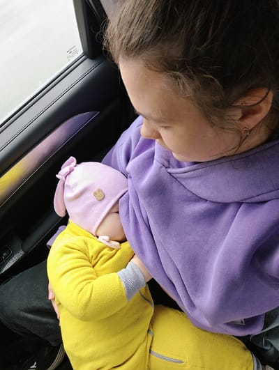 This vertical shot captures an intimate moment inside a car, likely in Kvarsa, Russia, where a mother is breastfeeding her infant. The mother, positioned on the right, has dark hair pulled back and wears a purple hooded sweatshirt. Her face is seen in profile, looking down at the baby with a gentle expression. A small earring is visible in her right ear.

The infant, held in the mother's arms, is dressed in a bright yellow fleece full-body outfit with grey cuffs. A light pink beanie adorned with bunny ears and a small brown bear-shaped patch covers the baby's head. The baby's face is largely obscured by the hat and the mother's clothing as it nurses. The mother's hand, with pink nail polish, is visible supporting the baby.

The setting is the interior of a car, with dark door panels featuring visible stitching. A window on the left reveals a bright, diffused light outside, suggesting an overcast or cloudy daytime. Text visible on the car window includes "HAVAL," "PREMO," "E4," and "43R-01030," which are likely manufacturer's marks or car specifications. A subtle purple reflection can be seen on the car door panel, possibly from the mother's attire or ambient lighting. The scene conveys a sense of nurturing and calm.