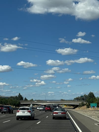 A view from a vehicle on a multi-lane highway in Citrus Heights, California, United States of America, shows traffic moving under a clear blue sky with scattered white clouds. Several cars are visible on the highway, including a white Honda CR-V in the foreground and a silver sedan in the middle lane. Overhead power lines stretch across the sky. A highway overpass is visible in the distance, with a sign indicating directions to "Roseville, Riverside Ave, Auburn Blvd". Trees and hills line the sides of the highway.